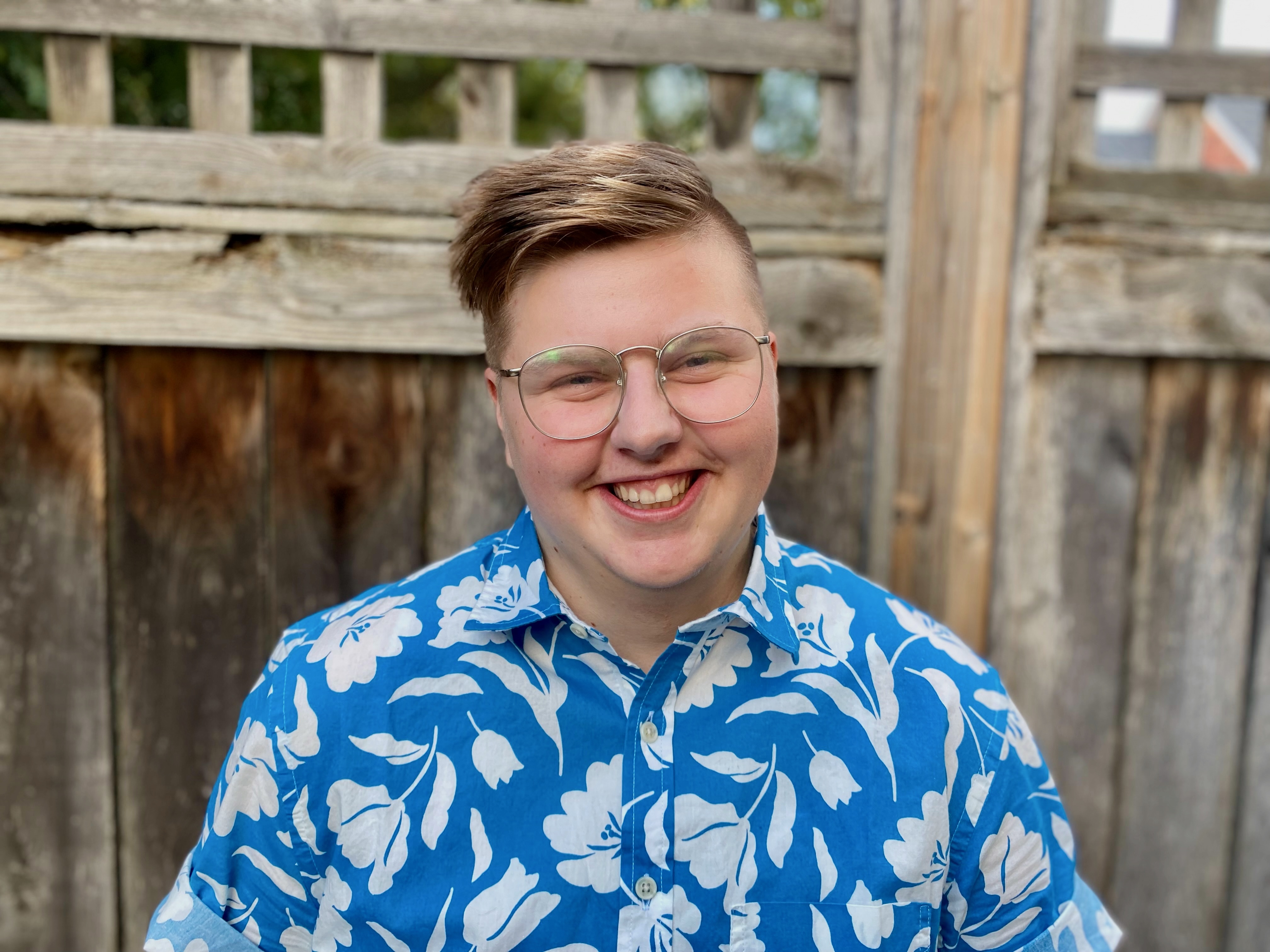 White transmasculine person with short blonde hair and glasses wearing a blue patterned button-down