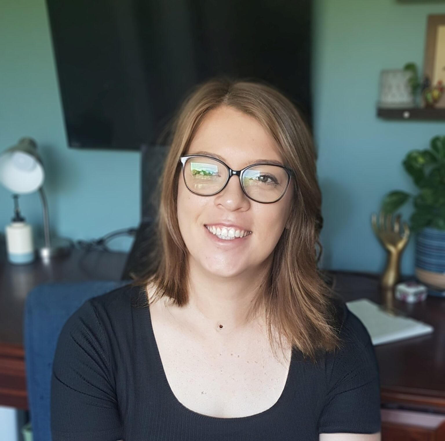 A white woman with brown hair and glasses sitting in a computer chair, smiling.